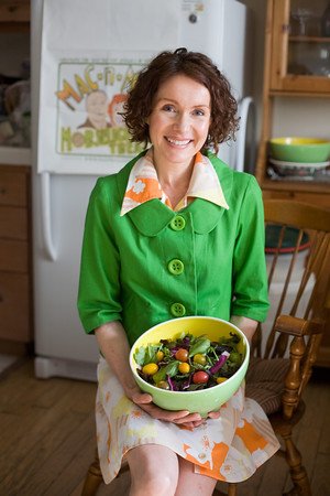 Certified Health Coach, Mo The Morselist, is sitting in a chair in a kitchen in an orange and yellow dress beneath a bright green jacket and posing with a green bowl of a rainbow of vegetables.